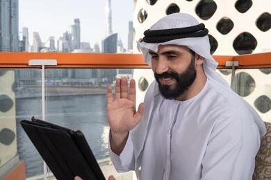 Gestures of joy and happiness, browsing the internet and using modern technologies, raising the hand upwards with gestures of greeting and peace, making a video call via a tablet device, working remotely through the internet, an Emirati Gulf Arab man wearing a kandura and a white ghutrah sitting in a high café overlooking the city.