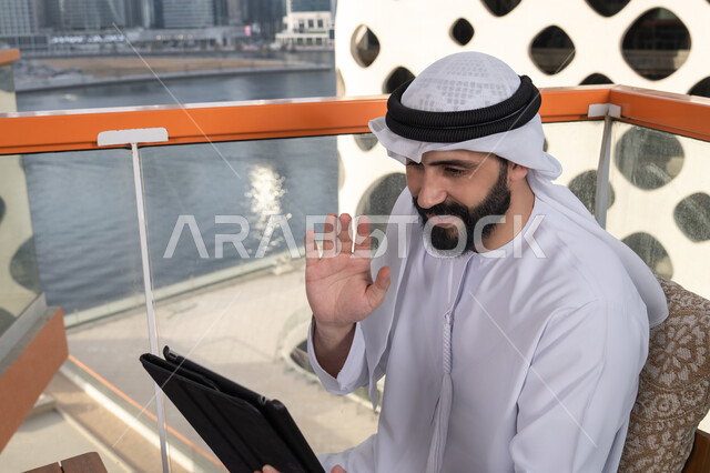 رفع الكف للأعلى بايماءات التحية والسلام, تصفح الانترنت واستخدام تقنيات حديثة, إجراء مكالمة فيديو عبر جهاز التابلت, ايماءات السرور والسعادة, العمل عن بعد من خلال الانترنت, رجل عربي خليجي اماراتي يرتدي الكندورة و الغتره البيضاء يجلس في مقهى مرتفع مطل على المدينة