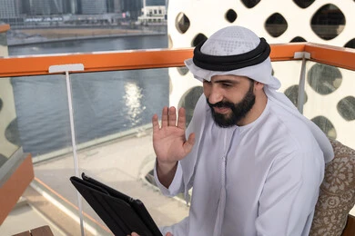 Raising the hand upwards with gestures of greeting and peace, browsing the internet and using modern technologies, making a video call via a tablet, gestures of joy and happiness, working remotely through the internet, an Emirati Gulf Arab man wearing a kandura and a white ghutrah sitting in a high café overlooking the city.