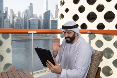 تصفح الانترنت واستخدام تقنيات حديثة, رفع الكف للأعلى بايماءات التحية والسلام, إجراء مكالمة فيديو عبر جهاز التابلت, ايماءات السرور والسعادة, العمل عن بعد من خلال الانترنت, رجل عربية خليجي اماراتي يرتدي الكندورة و الغتره البيضاء يجلس في مقهى مرتفع مطل على المدينة