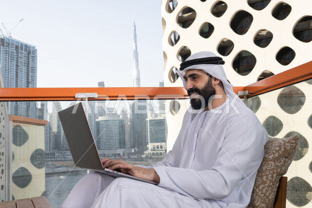 قضاء وقت ممتع بالخارج, الكتابة على لوحة المفاتيح، استخدام الأجهزة التقنية الحديثة والمتطورة لانجاز المشاريع في الكافيه، العمل عن بعد، رجل عربي خليجي اماراتي مبتسم يرتدي الكندورة والغترة البيضاء يجلس في المقهى ويستخدم جهاز الحاسوب المحمول بايماءات السرور