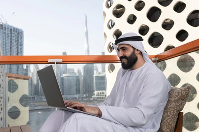 قضاء وقت ممتع بالخارج, الكتابة على لوحة المفاتيح، استخدام الأجهزة التقنية الحديثة والمتطورة لانجاز المشاريع في الكافيه، العمل عن بعد، رجل عربي خليجي اماراتي مبتسم يرتدي الكندورة والغترة البيضاء يجلس في المقهى ويستخدم جهاز الحاسوب المحمول بايماءات السرور