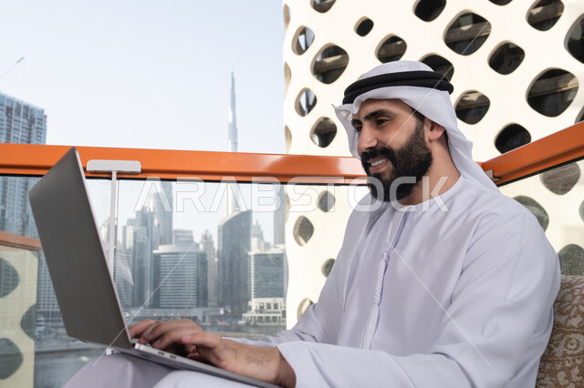 العمل لتحقيق الاهداف والاستراتيجيات, الكتابة على لوحة المفاتيح، استخدام الأجهزة التقنية الحديثة والمتطورة لانجاز المشاريع في الكافيه، العمل عن بعد، رجل عربي خليجي اماراتي مبتسم يرتدي الكندورة والغترة البيضاء يجلس في المقهى ويستخدم جهاز الحاسوب المحمول بايماءات السرور