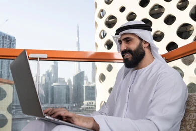 Working to achieve goals and strategies, typing on the keyboard, using modern and advanced technical devices to complete projects in the café, working remotely, a smiling Emirati Gulf Arab man wearing a kandura and a white ghutrah sitting in the café and using a laptop with gestures of joy.