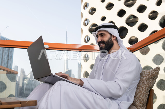 دمج العمل مع التقنية والتكنولوجيا, الكتابة على لوحة المفاتيح، العمل عن بعد، رجل عربي خليجي اماراتي مبتسم يرتدي الكندورة والغترة البيضاء يجلس في المقهى ويستخدم جهاز الحاسوب المحمول بايماءات السرور