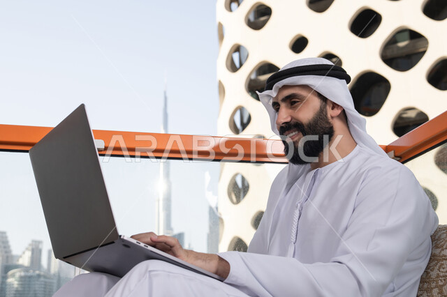 Completing remote work tasks, typing on the keyboard, using modern and advanced technological devices to accomplish projects in the café, a smiling Emirati Gulf Arab man wearing a kandura and a white ghutrah sits in the café using a laptop with gestures of joy.