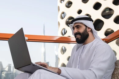إنجاز مهام العمل عن بعد،الكتابة على لوحة المفاتيح، استخدام الأجهزة التقنية الحديثة والمتطورة لانجاز المشاريع في الكافيه، رجل عربي خليجي اماراتي مبتسم يرتدي الكندورة والغترة البيضاء يجلس في المقهى ويستخدم جهاز الحاسوب المحمول بايماءات السرور
