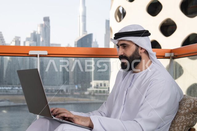 إدارة شؤون الأعمال وتنظيمها عبر الأجهزة التقنية الحديثة، الكتابة على لوحة المفاتيح، العمل عن بعد، رجل عربي خليجي اماراتي مبتسم يرتدي الكندورة والغترة البيضاء يجلس في المقهى ويستخدم جهاز الحاسوب المحمول بايماءات السرور