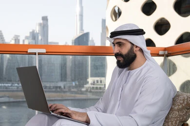 Managing and organizing business affairs through modern technological devices, typing on a keyboard, working remotely, a smiling Emirati Gulf Arab man wearing a kandura and a white ghutrah sitting in a café and using a laptop with gestures of joy.