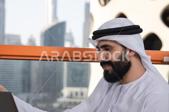 استخدام الأجهزة التقنية الحديثة والمتطورة لانجاز المشاريع في الكافيه, الكتابة على لوحة المفاتيح، العمل عن بعد، صورة مقربة لرجل عربي خليجي اماراتي مبتسم يرتدي الكندورة والغترة البيضاء يجلس في المقهى ويستخدم جهاز الحاسوب المحمول بايماءات السرور