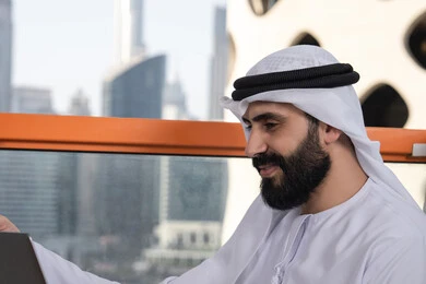 استخدام الأجهزة التقنية الحديثة والمتطورة لانجاز المشاريع في الكافيه, الكتابة على لوحة المفاتيح، العمل عن بعد، صورة مقربة لرجل عربي خليجي اماراتي مبتسم يرتدي الكندورة والغترة البيضاء يجلس في المقهى ويستخدم جهاز الحاسوب المحمول بايماءات السرور