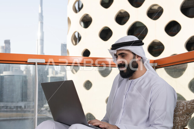 Typing on the keyboard, using modern and advanced technical devices to complete projects in the café, working remotely, a smiling Emirati Gulf Arab man wearing a kandura and a white ghutrah sitting in the café and using a laptop with gestures of joy.