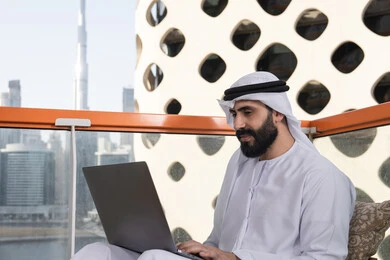 Typing on the keyboard, using modern and advanced technical devices to complete projects in the café, working remotely, a smiling Emirati Gulf Arab man wearing a kandura and a white ghutrah sitting in the café and using a laptop with gestures of joy.