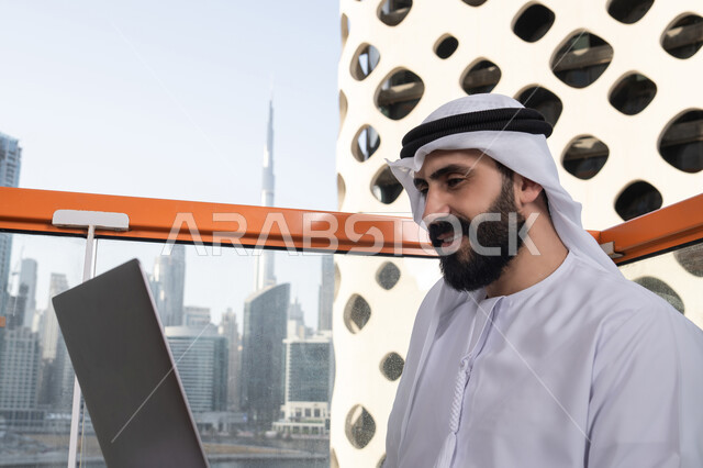 استخدام الأجهزة التقنية الحديثة والمتطورة لانجاز المشاريع في الكافيه، العمل عن بعد، صورة مقربة لرجل عربي خليجي اماراتي مبتسم يرتدي الكندورة والغترة البيضاء يجلس في المقهى ويستخدم جهاز الحاسوب المحمول بايماءات السرور