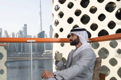 Spending free time enjoying a favorite drink, entertainment and leisure during the holiday, a side view of an Emirati Gulf Arab man wearing a kandura and a white ghutrah sitting in one of the cafes holding a cup of coffee, having a pleasant and entertaining time outdoors, cafes and restaurants in the United Arab Emirates.