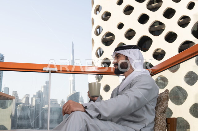 Enjoying favorite drinks, entertainment and leisure during the holiday, a side view of an Emirati Gulf Arab man wearing a kandura and a white ghutrah sitting in one of the cafes holding a cup of coffee, having a fun and enjoyable time outdoors, cafes and restaurants in the United Arab Emirates.