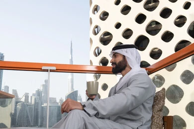 Enjoying favorite drinks, entertainment and leisure during the holiday, a side view of an Emirati Gulf Arab man wearing a kandura and a white ghutrah sitting in one of the cafes holding a cup of coffee, having a fun and enjoyable time outdoors, cafes and restaurants in the United Arab Emirates.