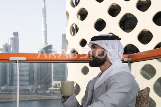 الكافيهات والمطاعم في الامارات العربية المتحدة, التسلية والترفيه في العطلة، صورة مقربة من الجانب لرجل عربي خليجي اماراتي يرتدي الكندورة والغترة البيضاء يجلس فى احدي المقاهي و يمسك بيده كوب من القهوه، قضاء وقت ممتع ومسلي في الخارج