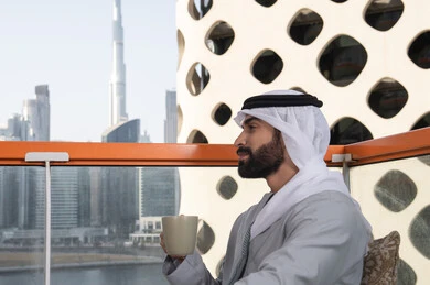 الكافيهات والمطاعم في الامارات العربية المتحدة, التسلية والترفيه في العطلة، صورة مقربة من الجانب لرجل عربي خليجي اماراتي يرتدي الكندورة والغترة البيضاء يجلس فى احدي المقاهي و يمسك بيده كوب من القهوه، قضاء وقت ممتع ومسلي في الخارج