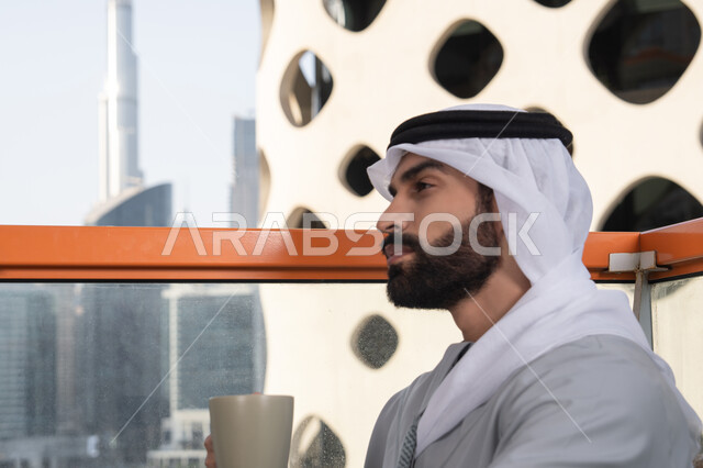 قضاء وقت ممتع ومسلي في الخارج, التسلية والترفيه في العطلة, صورة من الجانب لرجل عربي خليجي اماراتي يرتدي الكندورة والغترة البيضاء يجلس فى احدي المقاهي و يمسك بيده كوب من القهوه، الكافيهات والمطاعم في الامارات العربية المتحدة