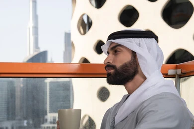 قضاء وقت ممتع ومسلي في الخارج, التسلية والترفيه في العطلة, صورة من الجانب لرجل عربي خليجي اماراتي يرتدي الكندورة والغترة البيضاء يجلس فى احدي المقاهي و يمسك بيده كوب من القهوه، الكافيهات والمطاعم في الامارات العربية المتحدة