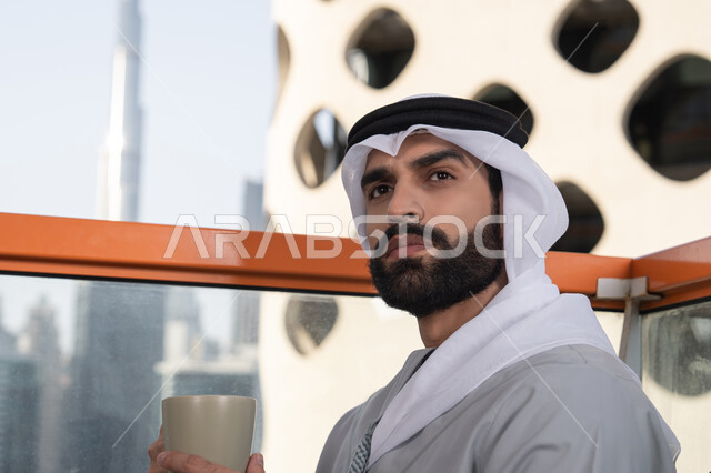 التسلية والترفيه في العطلة،  صورة مقربة لرجل عربي خليجي اماراتي يرتدي الكندورة والغترة البيضاء يجلس فى احدي المقاهي و يمسك بيده كوب من القهوه ينظر الى شيء ما، قضاء وقت ممتع ومسلي في الخارج، الكافيهات والمطاعم في الامارات العربية المتحدة