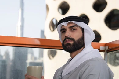 التسلية والترفيه في العطلة،  صورة مقربة لرجل عربي خليجي اماراتي يرتدي الكندورة والغترة البيضاء يجلس فى احدي المقاهي و يمسك بيده كوب من القهوه ينظر الى شيء ما، قضاء وقت ممتع ومسلي في الخارج، الكافيهات والمطاعم في الامارات العربية المتحدة