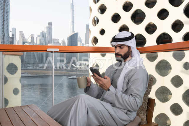 تناول المشروبات المفضله, تبادل الأحاديث والتواصل والأخبار مع الأهل والأصدقاء، رجل عربي خليجي اماراتي يرتدي الكندورة والغترة البيضاء يمسك بيده الهاتف المحمول و فنجان من القهوه، استخدام جهاز تقني حديث ومتطور، العمل عن بعد، تصفح التطبيقات وبرامج التواصل الاجتماعي