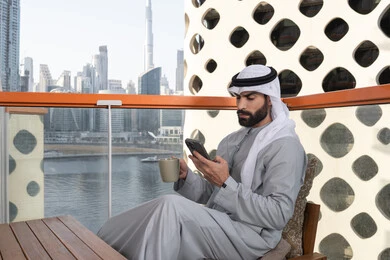 تناول المشروبات المفضله, تبادل الأحاديث والتواصل والأخبار مع الأهل والأصدقاء، رجل عربي خليجي اماراتي يرتدي الكندورة والغترة البيضاء يمسك بيده الهاتف المحمول و فنجان من القهوه، استخدام جهاز تقني حديث ومتطور، العمل عن بعد، تصفح التطبيقات وبرامج التواصل الاجتماعي