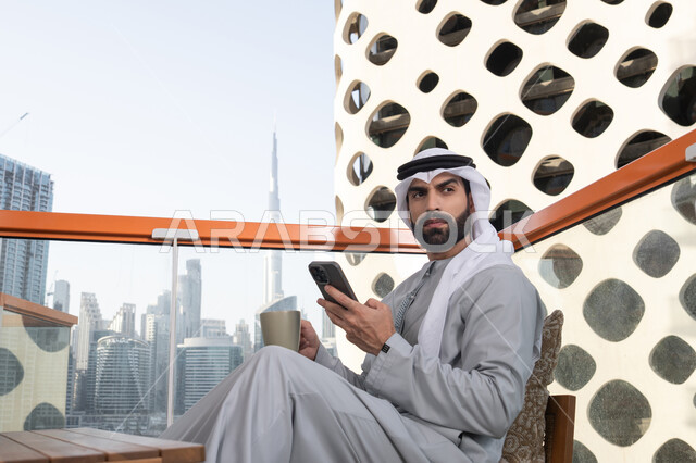 الاستمتاع بقضاء وقت ممتع فى الخارج, تبادل الأحاديث والتواصل والأخبار مع الأهل والأصدقاء، رجل عربي خليجي اماراتي يرتدي الكندورة والغترة البيضاء يمسك بيده الهاتف المحمول و ينظر الى شيء ما، استخدام جهاز تقني حديث ومتطور، تناول المشروبات المفضله، تصفح التطبيقات وبرامج التواصل الاجتماعي