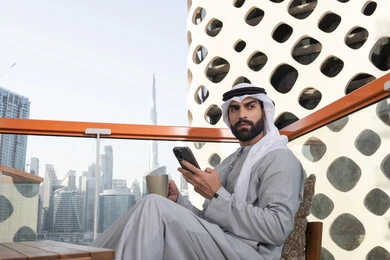 Enjoying a good time outdoors, exchanging conversations and news with family and friends, an Arab Gulf Emirati man wearing a kandura and a white ghutrah holding a mobile phone and looking at something, using a modern and advanced technological device, enjoying favorite drinks, browsing applications and social media programs.