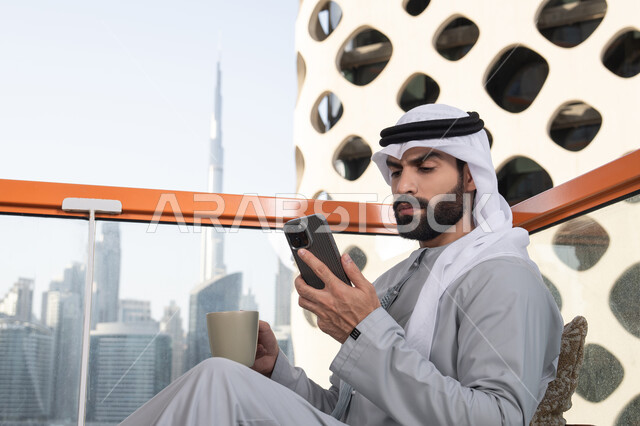 Browsing applications and social media programs, working remotely, exchanging conversations and communication and news with family and friends, an Arab Gulf Emirati man wearing a kandura and a white ghutrah holding a mobile phone in his hand, using a modern and advanced technical device, enjoying his favorite drink.