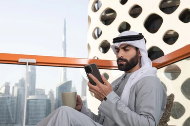 تصفح التطبيقات وبرامج التواصل الاجتماعي, العمل عن بعد, تبادل الأحاديث والتواصل والأخبار مع الأهل والأصدقاء، رجل عربي خليجي اماراتي يرتدي الكندورة والغترة البيضاء يمسك بيده الهاتف المحمول، استخدام جهاز تقني حديث ومتطور, تناول المشروب المفضل