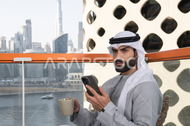 Remote work, exchanging conversations and communication and news with family and friends, an Arab Gulf Emirati man wearing a kandura and a white ghutrah holding a mobile phone in his hand, using a modern and advanced technical device, browsing applications and social media programs, enjoying his favorite drink.