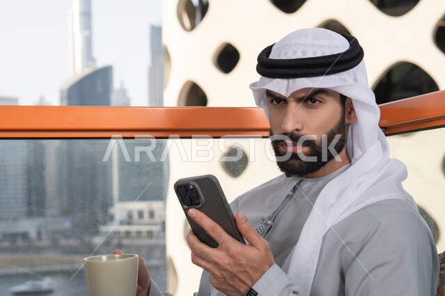 استخدام جهاز تقني حديث ومتطور, تبادل الأحاديث والتواصل والأخبار مع الأهل والأصدقاء، رجل عربي خليجي اماراتي يرتدي الكندورة والغترة البيضاء يمسك بيده الهاتف المحمول، العمل عن بعد، تصفح التطبيقات وبرامج التواصل الاجتماعي, تناول المشروب المفضل