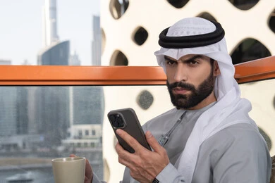 استخدام جهاز تقني حديث ومتطور, تبادل الأحاديث والتواصل والأخبار مع الأهل والأصدقاء، رجل عربي خليجي اماراتي يرتدي الكندورة والغترة البيضاء يمسك بيده الهاتف المحمول، العمل عن بعد، تصفح التطبيقات وبرامج التواصل الاجتماعي, تناول المشروب المفضل
