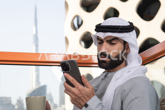 استخدام جهاز تقني حديث ومتطور, تبادل الأحاديث والتواصل والأخبار مع الأهل والأصدقاء، رجل عربي خليجي اماراتي يرتدي الكندورة والغترة البيضاء يمسك بيده الهاتف المحمول، العمل عن بعد، تصفح التطبيقات وبرامج التواصل الاجتماعي, تناول المشروب المفضل