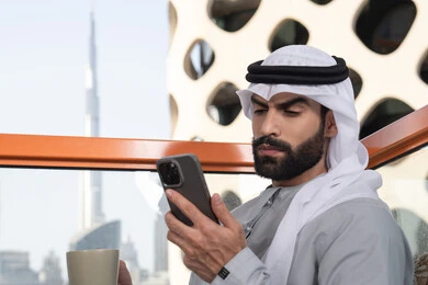 استخدام جهاز تقني حديث ومتطور, تبادل الأحاديث والتواصل والأخبار مع الأهل والأصدقاء، رجل عربي خليجي اماراتي يرتدي الكندورة والغترة البيضاء يمسك بيده الهاتف المحمول، العمل عن بعد، تصفح التطبيقات وبرامج التواصل الاجتماعي, تناول المشروب المفضل