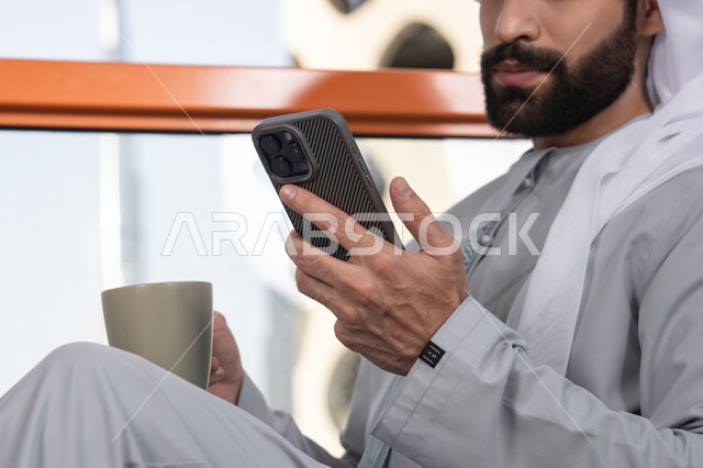 رجل عربي خليجي اماراتي يرتدي الكندورة والغترة البيضاء يمسك بيده الهاتف المحمول, تبادل الأحاديث والتواصل والأخبار مع الأهل والأصدقاء، استخدام جهاز تقني حديث ومتطور، العمل عن بعد، تصفح التطبيقات وبرامج التواصل الاجتماعي, تناول المشروب المفضل