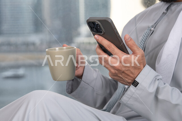 Exchanging conversations, communication, and news with family and friends, a close-up image of the hand of an Emirati Arab man wearing a kandura holding a mobile phone, using a modern and advanced technological device, working remotely, browsing applications and social media programs, enjoying a favorite drink.