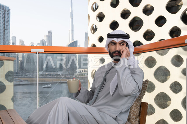 Chatting and conversing using a modern technical device, making a phone call, sipping a favorite drink, an Emirati Gulf Arab man wearing a kandura and a white ghutrah holding a mobile phone and a cup of coffee, working remotely, communicating and exchanging conversations with family and friends.