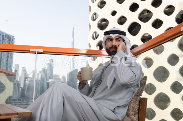 Integrating modern technologies and technology into practical life, making a phone call, sipping a favorite drink, an Arab Gulf Emirati man wearing a kandura and a white ghutrah holding a mobile phone and a cup of coffee, remote work, communicating and exchanging conversations with family and friends.