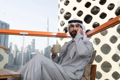 Integrating modern technologies and technology into practical life, making a phone call, sipping a favorite drink, an Arab Gulf Emirati man wearing a kandura and a white ghutrah holding a mobile phone and a cup of coffee, remote work, communicating and exchanging conversations with family and friends.