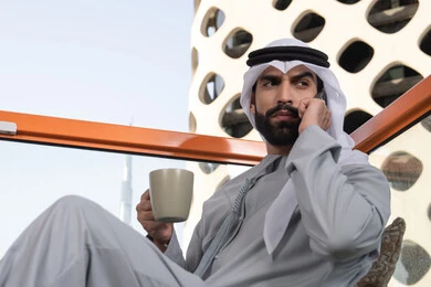 قضاء عطله نهاية الاسبوع بالخارج, اجراء مكالمة هاتفية، استخدام جهاز تقني حديث ومتطور، احتساء المشروب المفضل، رجل عربي خليجي اماراتي يرتدي الكندورة والغترة البيضاء يمسك بيده الهاتف المحمول وكوب من القهوة، العمل عن بعد، التواصل وتبادل الأحاديث مع الأهل والأصدقاء