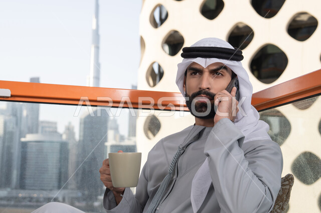 الاستمتاع بتناول المشروبات, اجراء مكالمة هاتفية، استخدام جهاز تقني حديث ومتطور، رجل عربي خليجي اماراتي يرتدي الكندورة والغترة البيضاء يمسك بيده الهاتف المحمول وكوب من القهوة، العمل عن بعد، التواصل وتبادل الأحاديث مع الأهل والأصدقاء