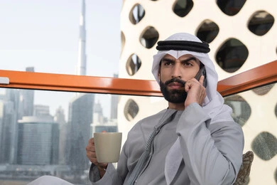 Enjoying drinks, making a phone call, using a modern and advanced technological device, an Emirati Gulf Arab man wearing a kandura and a white ghutrah holding a mobile phone and a cup of coffee, working remotely, communicating and exchanging conversations with family and friends.