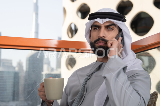 قضاء وقت ممتع فى الخارج. اجراء مكالمة هاتفية، استخدام جهاز تقني حديث ومتطور، احتساء المشروب المفضل، رجل عربي خليجي اماراتي يرتدي الكندورة والغترة البيضاء يمسك بيده الهاتف المحمول وكوب من القهوة، العمل عن بعد، التواصل وتبادل الأحاديث مع الأهل والأصدقاء