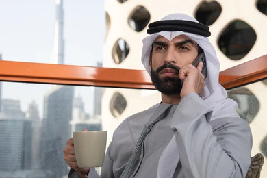 قضاء وقت ممتع فى الخارج. اجراء مكالمة هاتفية، استخدام جهاز تقني حديث ومتطور، احتساء المشروب المفضل، رجل عربي خليجي اماراتي يرتدي الكندورة والغترة البيضاء يمسك بيده الهاتف المحمول وكوب من القهوة، العمل عن بعد، التواصل وتبادل الأحاديث مع الأهل والأصدقاء