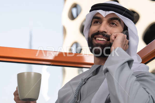 استخدام التكنولوجيا والتقنيات الحديثه، ايماءات السعادة و السرور, اجراء مكالمة هاتفية، احتساء المشروب المفضل، رجل عربي خليجي اماراتي يرتدي الكندورة والغترة البيضاء يمسك بيده الهاتف المحمول وكوب من القهوة، العمل عن بعد، التواصل وتبادل الأحاديث مع الأهل والأصدقاء