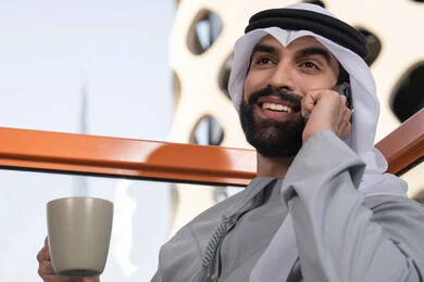 استخدام التكنولوجيا والتقنيات الحديثه، ايماءات السعادة و السرور, اجراء مكالمة هاتفية، احتساء المشروب المفضل، رجل عربي خليجي اماراتي يرتدي الكندورة والغترة البيضاء يمسك بيده الهاتف المحمول وكوب من القهوة، العمل عن بعد، التواصل وتبادل الأحاديث مع الأهل والأصدقاء